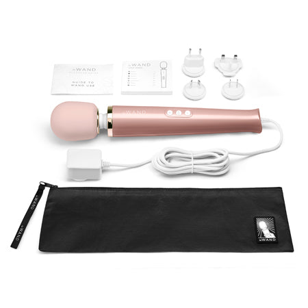 A rose gold Le Wand Plug-In Vibrating Massager with a sleek design, accompanied by a black carrying case, a USB charging cable, and an AC power adapter.