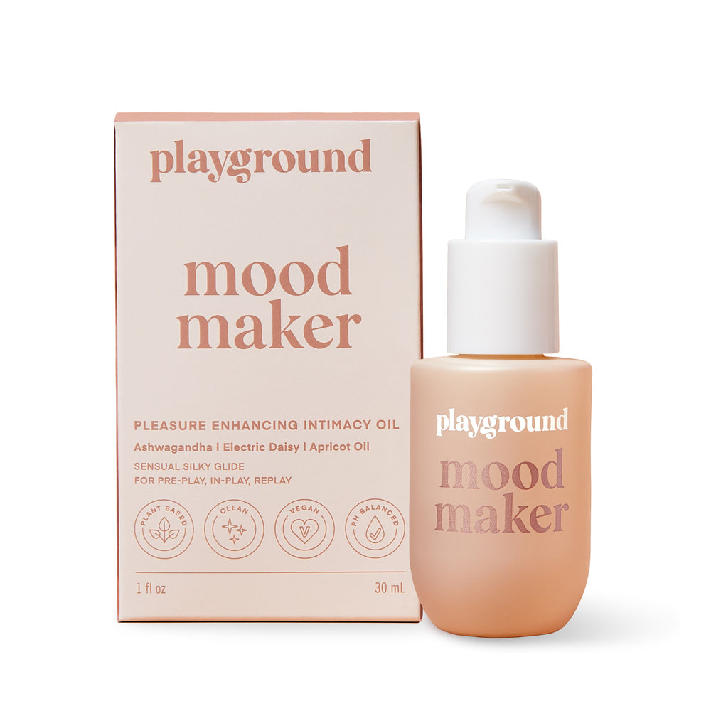 The image shows a product packaging and bottle for Playground Mood Maker Intimacy Oil. The packaging is pink with text and the bottle is transparent, showing the oil inside.
