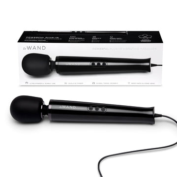 A black Le Wand Plug-In Vibrating Massager with a sleek design, showing the wand itself and the box in the background. The packaging highlights the product's features and includes a black silicone attachment.